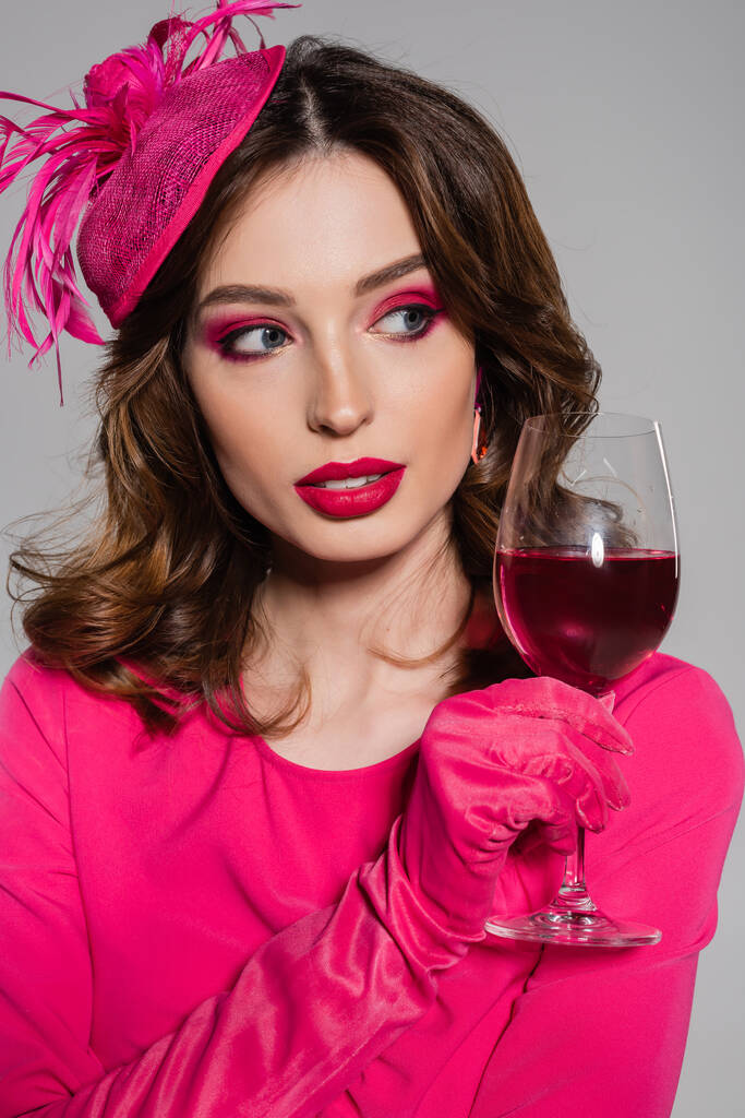 portrait of young woman in elegant hat and magenta color dress holding glass with alcohol drink isolated on grey  - Foto, Imagem