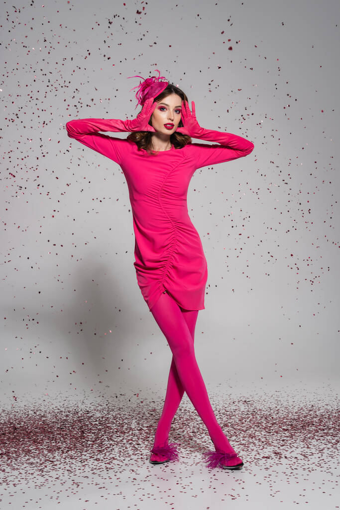 full length of brunette woman in elegant hat and magenta color dress posing on grey with falling confetti  - Foto, afbeelding