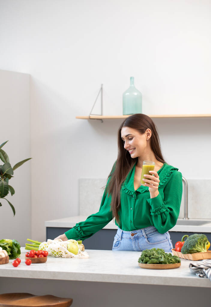 Una giovane bruna attraente tiene un bicchiere con un frullato verde, in piedi a un tavolo con verdure fresche e sorrisi. Il concetto di alimentazione sana, disintossicazione e dieta. - Foto, immagini