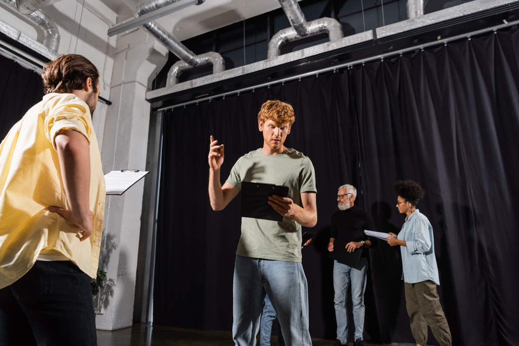 redhead man gesturing while looking at screenplay and rehearsing near multiethnic actors in theater - Fotoğraf, Görsel