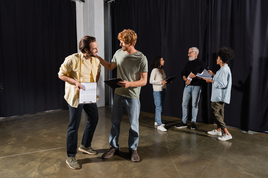 smiling men with scenarios talking in theater school near art director and interracial actors on background - Foto, imagen