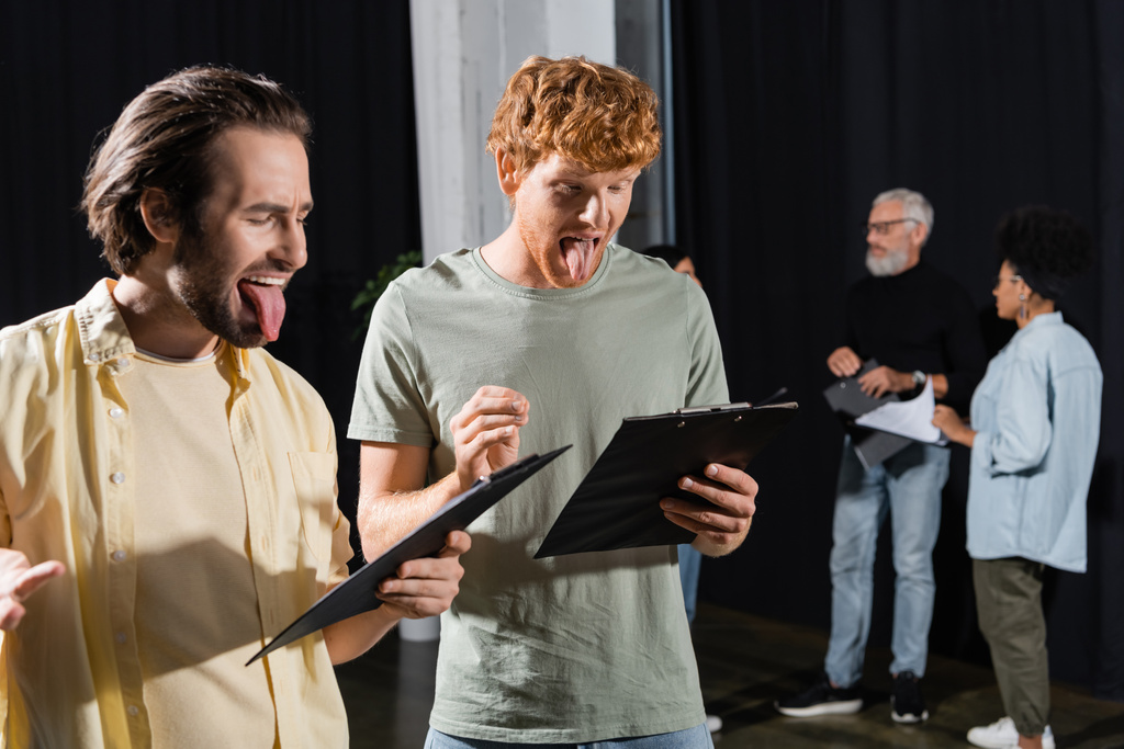brunette and redhead men sticking out tongues while reading scenarios during rehearsal in theater - Фото, изображение