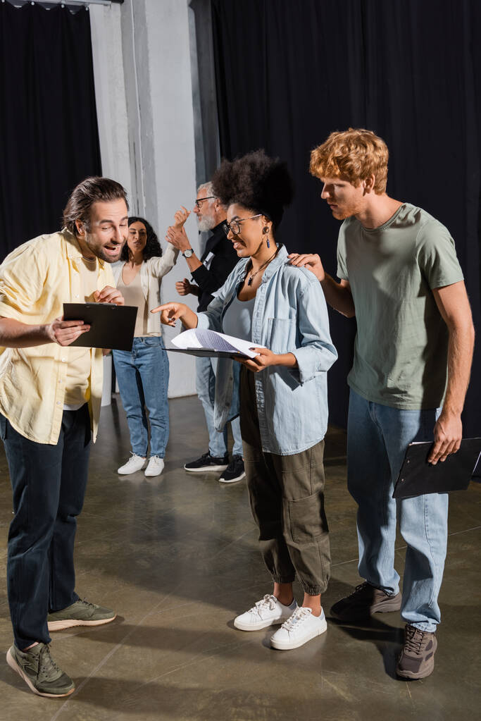 positive man showing clipboard with scenario to interracial actors on stage in theater - Foto, immagini