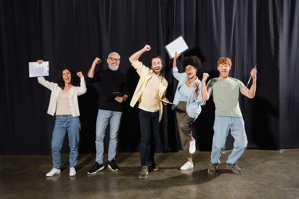 excited interracial actors with mature art director rejoicing on stage and showing triumph gesture. Translation of tattoo: om, shanti, peace - Foto, imagen