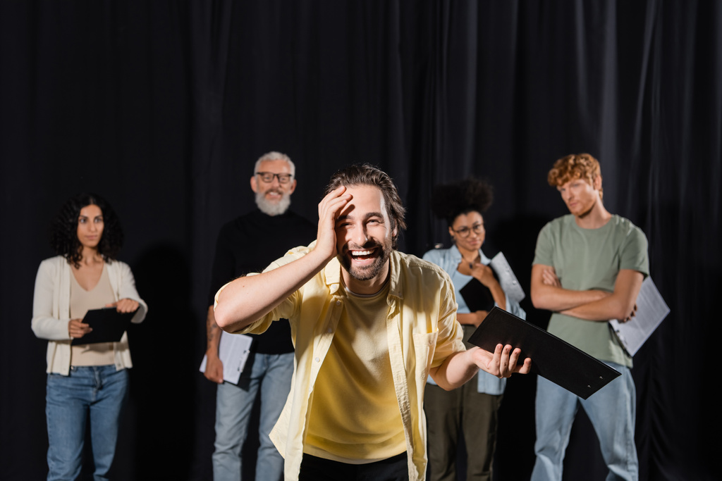 laughing man touching face and looking at camera near blurred multiethnic actors and producer in theater school - 写真・画像