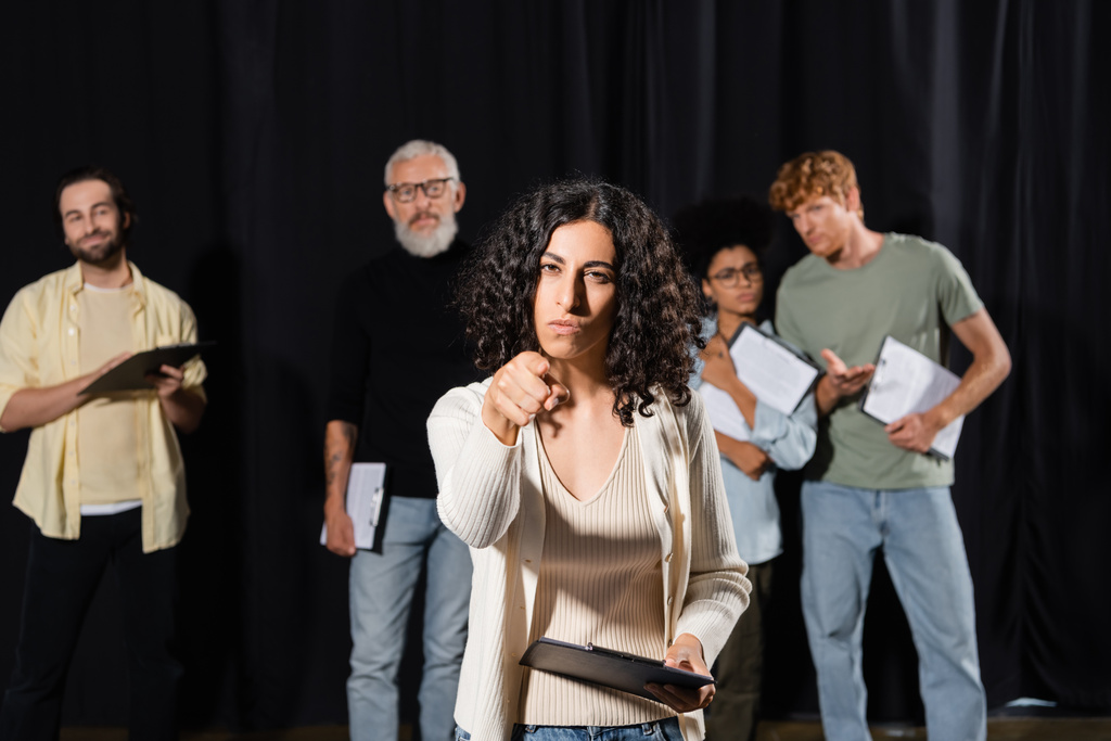 multiracial woman with serious face expression pointing at camera while rehearsing near blurred actors and mature art director - Fotoğraf, Görsel