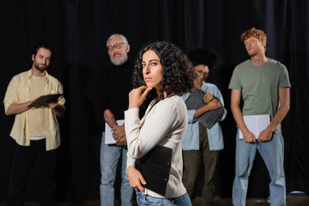 multiracial woman with strict face expression looking at camera while rehearsing near art director and actors on blurred background - Photo, Image