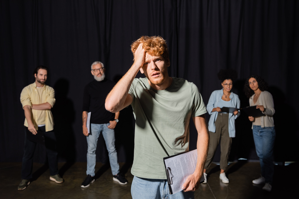 frustrated redhead man holding clipboard and touching head while looking at camera near actors and producer on blurred background - Foto, afbeelding