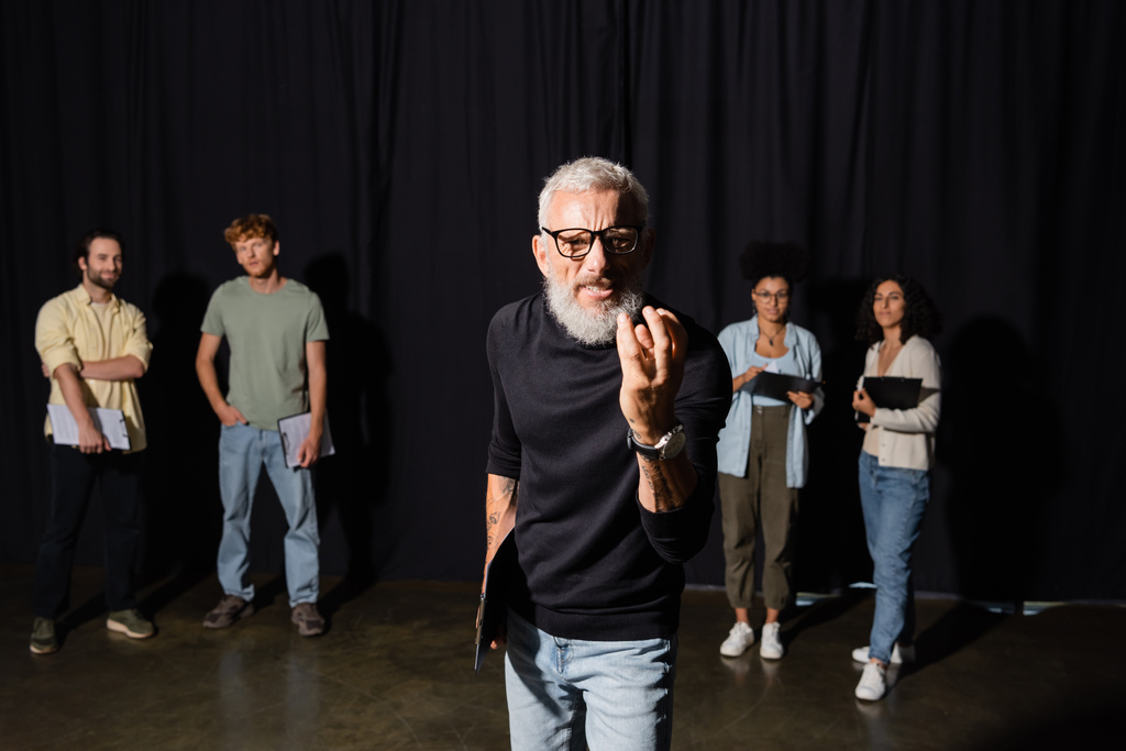bearded art director in eyeglasses looking at camera and gesturing near multiracial students on blurred background - Foto, Imagem
