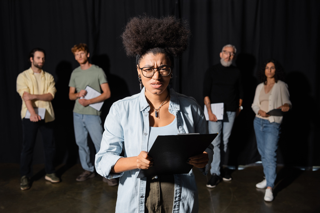frowning african american woman holding clipboard and looking at camera near multicultural actors and producer on blurred background - 写真・画像