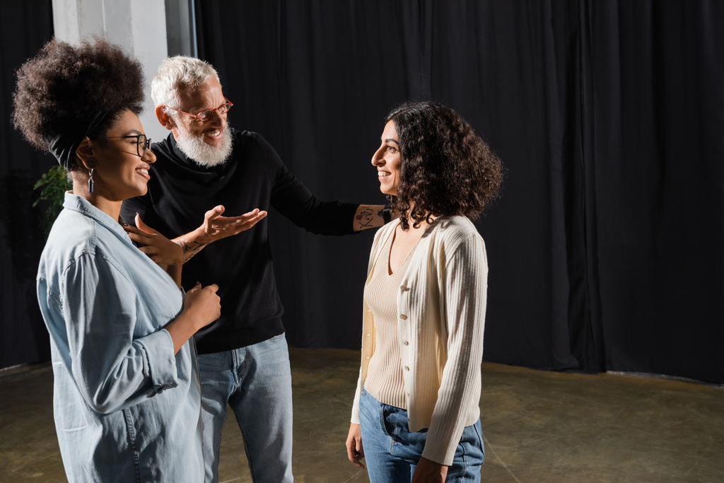 smiling art director pointing with hand and talking to multiracial woman near african american actress in theater - Photo, image