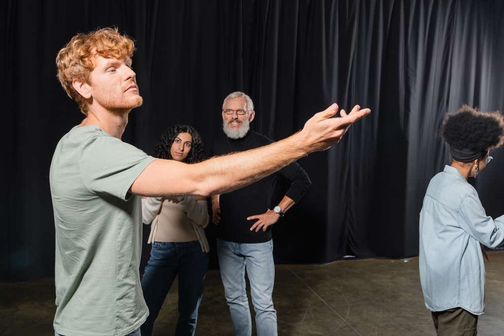 redhead man posing with outstretched hand near interracial women and smiling art director in theater - Fotografie, Obrázek