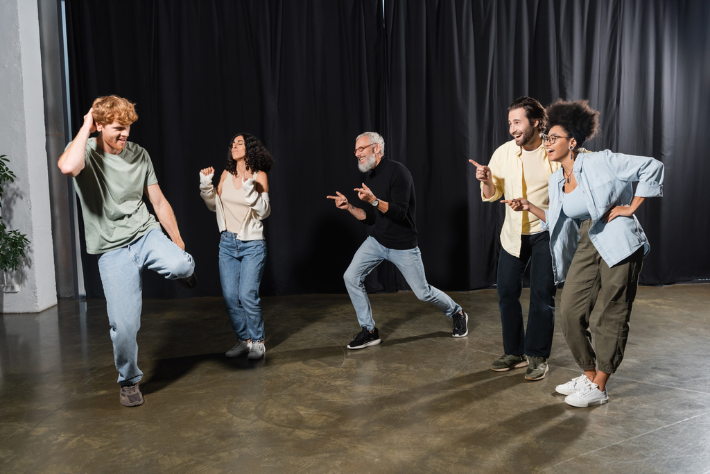 веселі мультикультурні актори, що вказують на репетиції рудого чоловіка на сцені театру
  - Фото, зображення