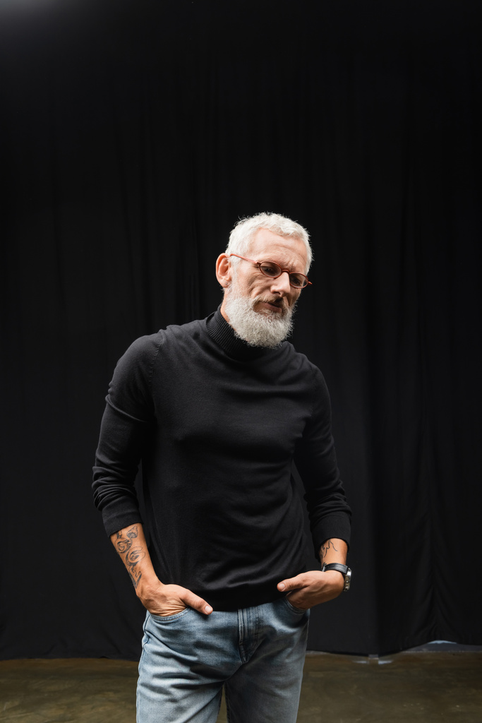 mature tattooed actor in eyeglasses and black turtleneck standing with hands in pockets of jeans in theater - Foto, Imagem