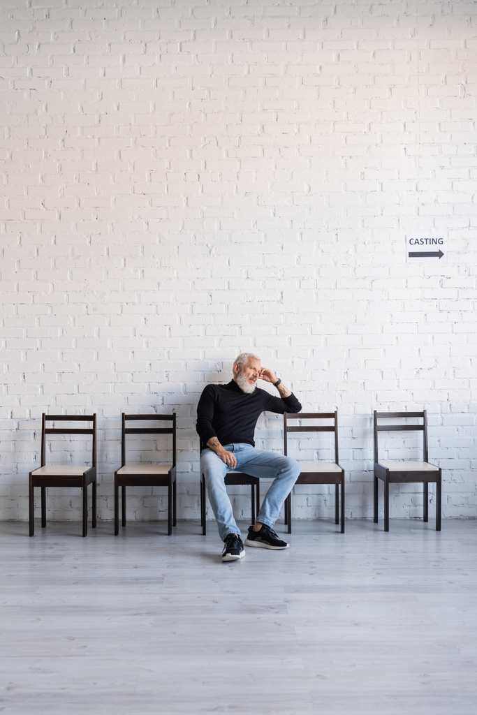 нудний сіроволосий чоловік сидить на стільці біля білої стіни і чекає на кастинг
 - Фото, зображення