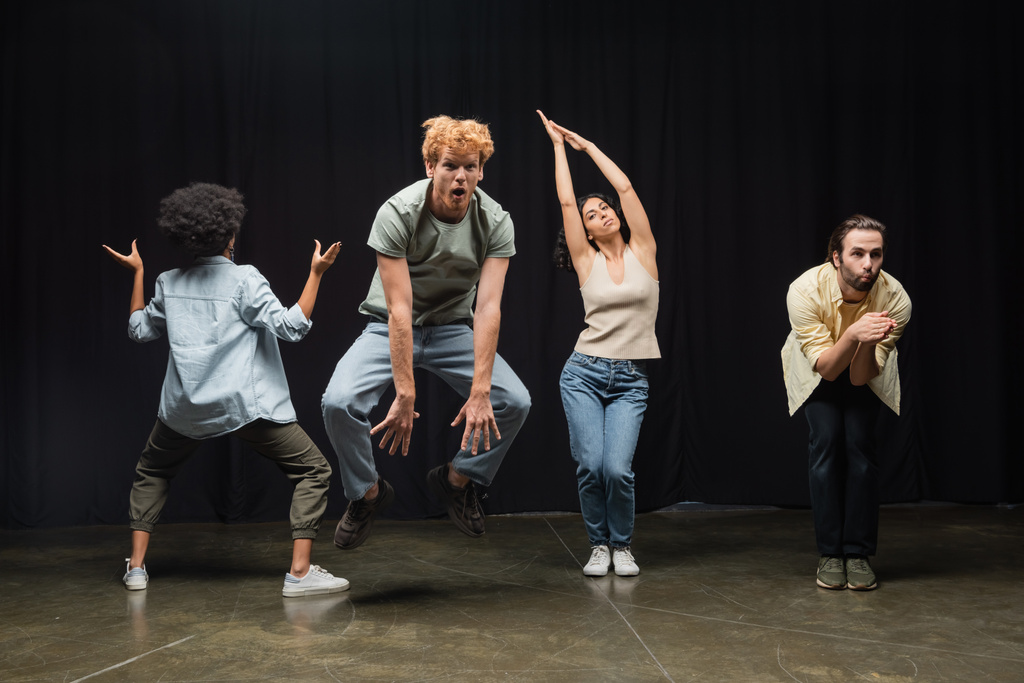 excited redhead man jumping near multiethnic actors posing during rehearsal in theater - Foto, immagini