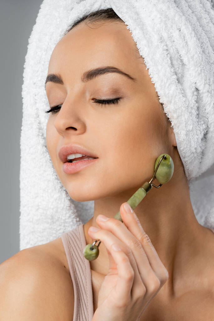 Portrait of young woman with towel on head using jade roller isolated on grey  - Photo, Image