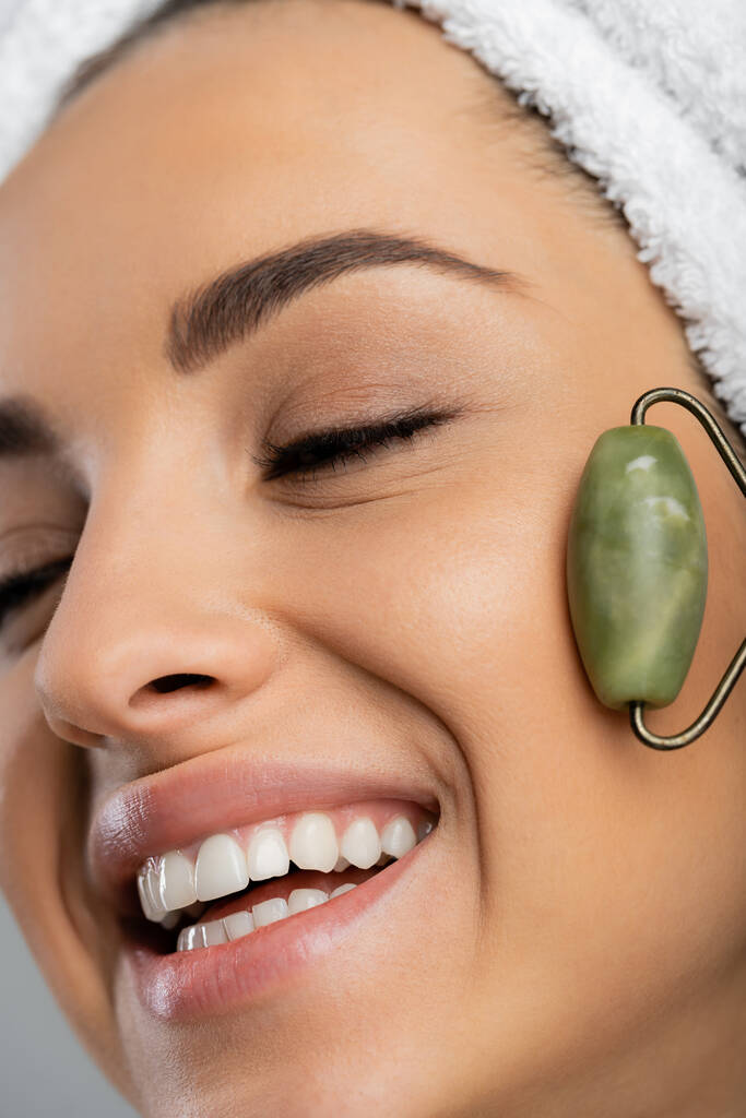 Close up view of smiling woman with towel on head massaging face with jade roller isolated on grey  - Fotó, kép