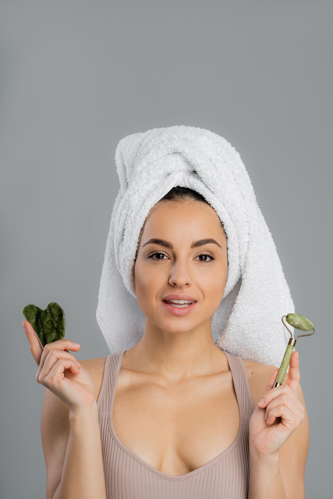 Young woman with towel on head holding jade roller and gua sha isolated on grey  - Foto, Bild