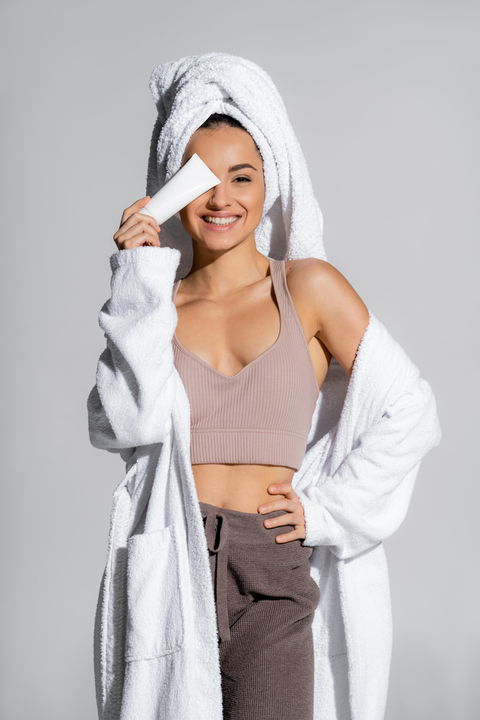 Cheerful woman in bathrobe holding body lotion near face isolated on grey  - 写真・画像