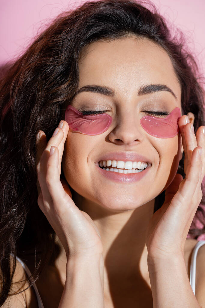 Curly woman touching hydrogel eye patches on face on pink background  - Фото, изображение