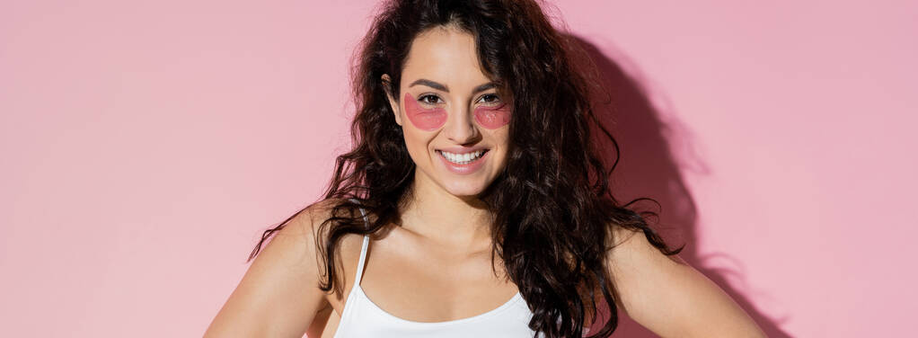 Smiling and curly woman with hydrogel eye patches looking at camera on pink background, banner  - Foto, immagini