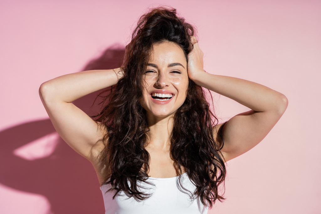 Cheerful freckled woman touching hair on pink background  - Photo, image