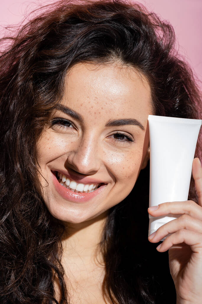 Cheerful young woman with freckles holding face cream isolated on pink  - Foto, Imagen