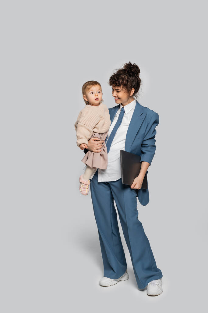 повна довжина весела бізнес-леді в синьому костюмі тримає в руках дочку малюка і ноутбук на сірому
  - Фото, зображення