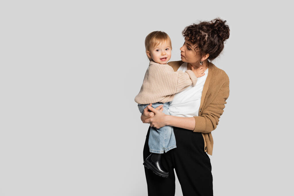 stylish mother in casual clothes holding in arms cheerful baby girl in sweater isolated on grey  - Fotoğraf, Görsel