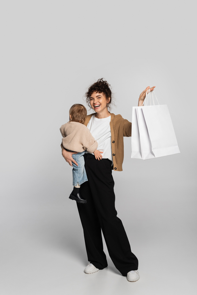 full length of cheerful mother in casual clothes holding in arms toddler baby and shopping bags on grey  - Foto, immagini