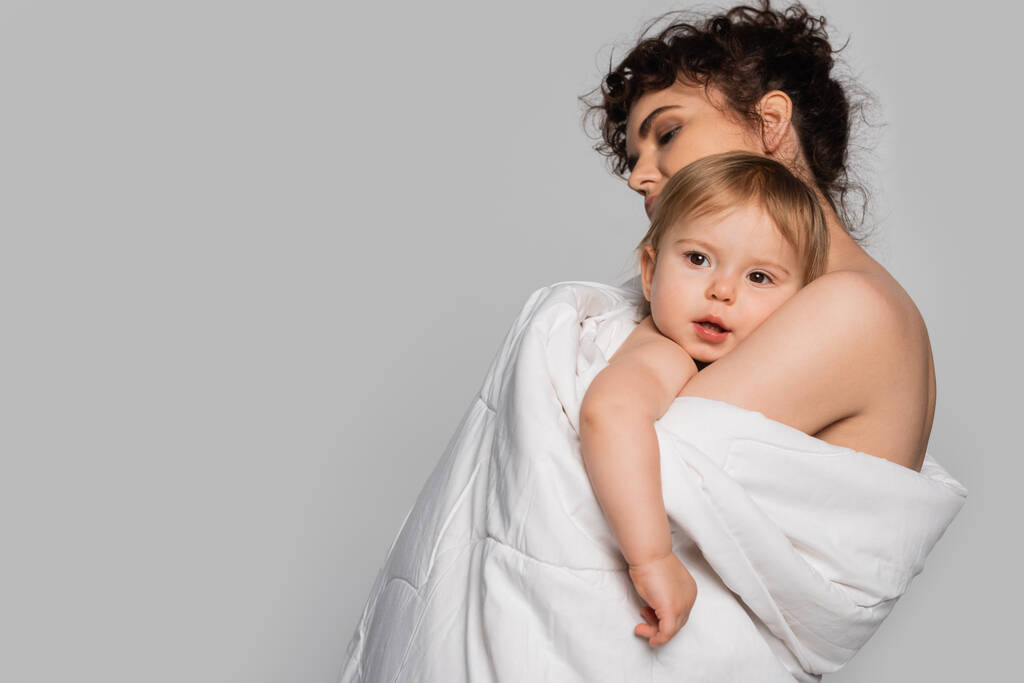 curly mother hugging toddler daughter covered in white duvet isolated on grey  - Foto, Imagem
