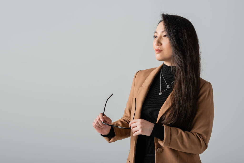 asian businesswoman in beige blazer holding glasses and looking away isolated on grey  - Foto, afbeelding