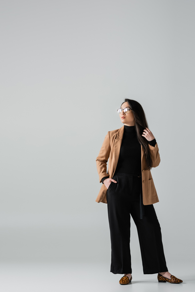 повна довжина впевненої азіатської бізнес-леді в бежевих блейзер і чорних штанях, що стоять з рукою в кишені на сірому
  - Фото, зображення