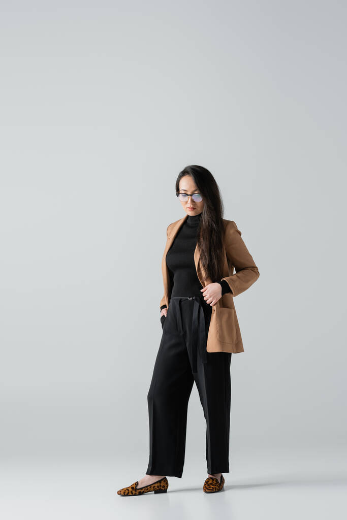 full length of asian businesswoman in beige blazer and black pants standing with hand in pocket on grey  - Fotó, kép