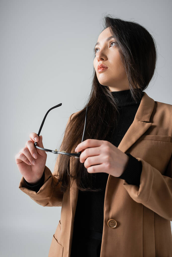 low angle view of asian businesswoman in beige blazer and black turtleneck holding glasses isolated on grey  - Valokuva, kuva
