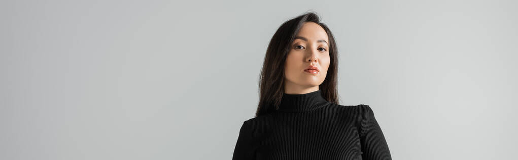 brunette asian businesswoman in black turtleneck looking at camera isolated on grey, banner  - Foto, imagen