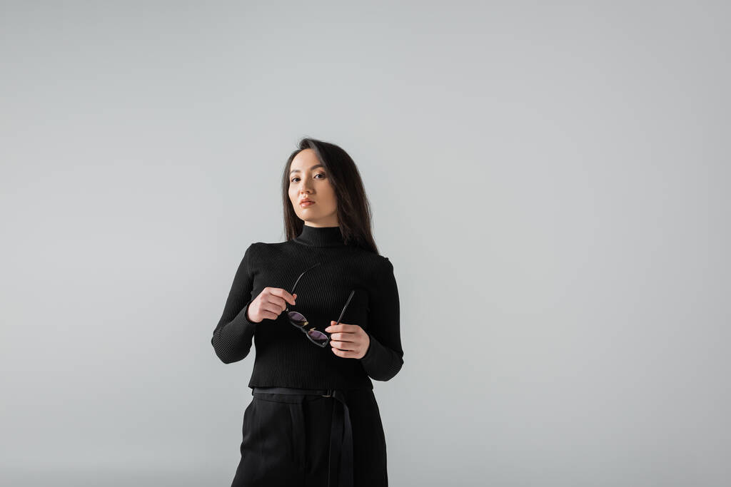 brunette asian businesswoman in black turtleneck looking at camera while holding glasses isolated on grey  - Foto, Imagem