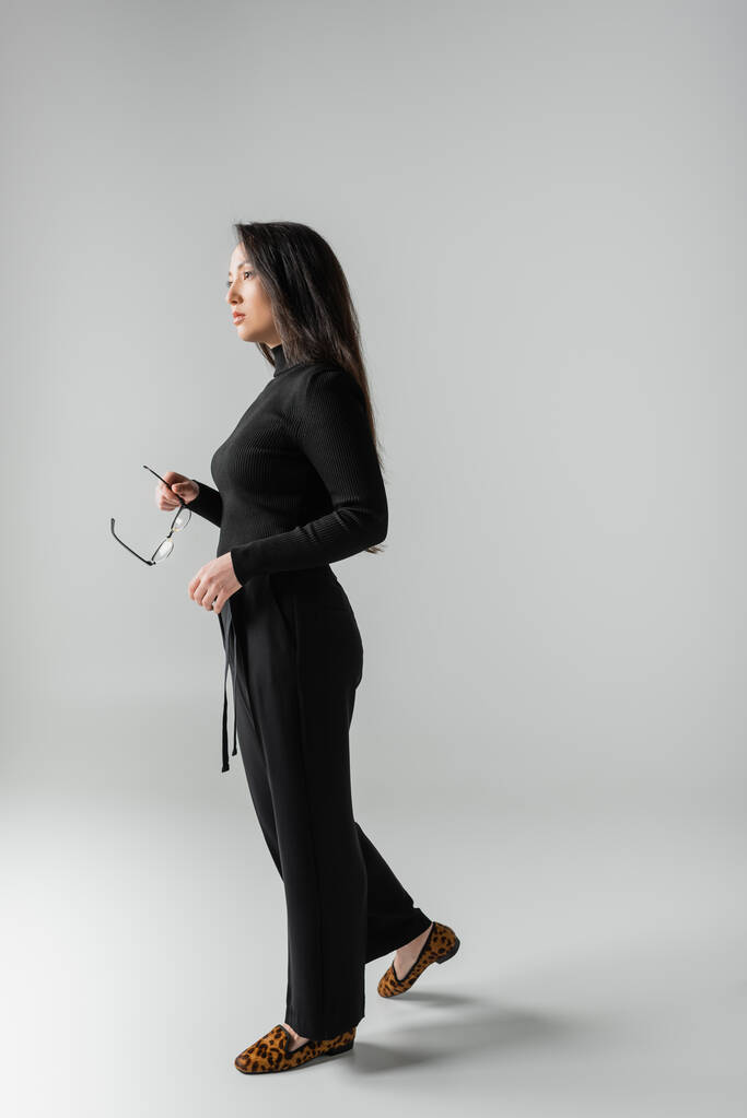 full length of brunette asian woman in black outfit and loafers with animal print holding glasses on grey  - Fotografie, Obrázek