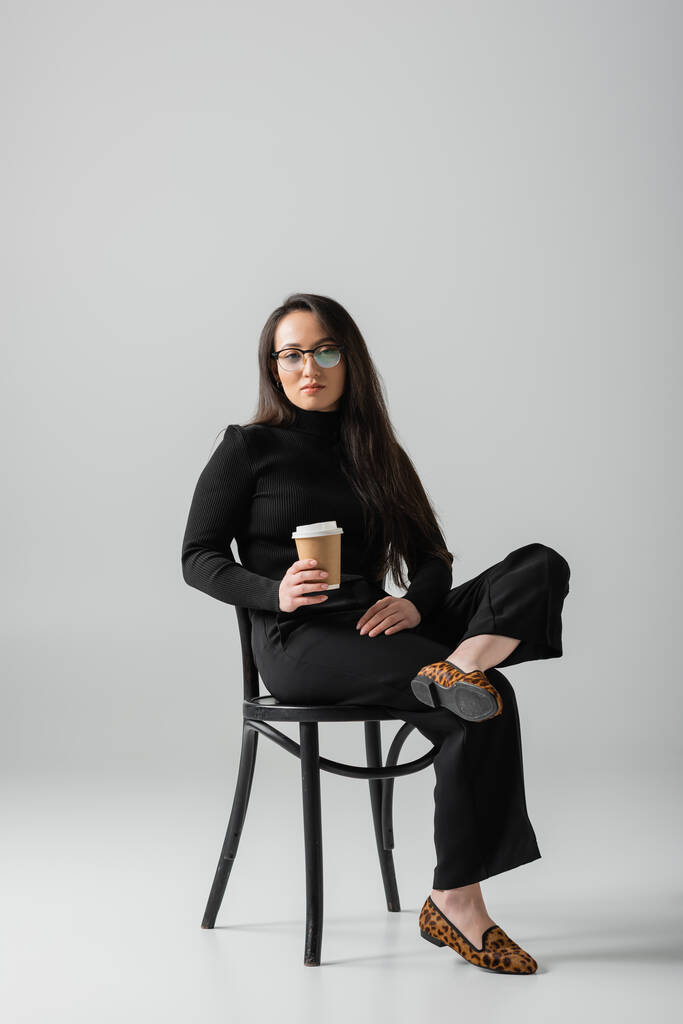 full length of asian woman in black outfit and glasses holding paper cup while sitting on chair on grey  - Photo, Image