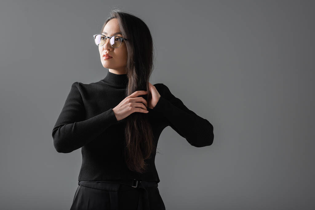 portrait of brunette and asian woman in black turtleneck and glasses looking away isolated on dark grey  - Photo, Image