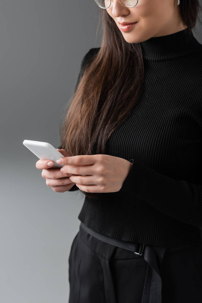 cropped view of young woman in black turtleneck and glasses using smartphone on dark grey  - Photo, Image
