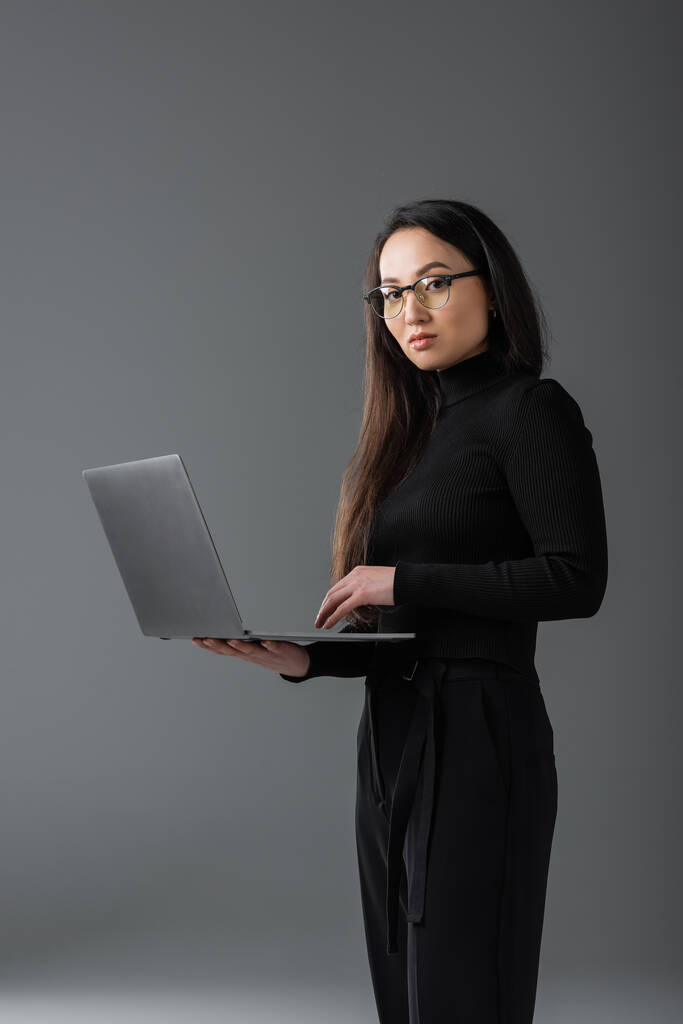 pretty asian freelancer in black turtleneck and glasses holding laptop on dark grey  - Foto, immagini