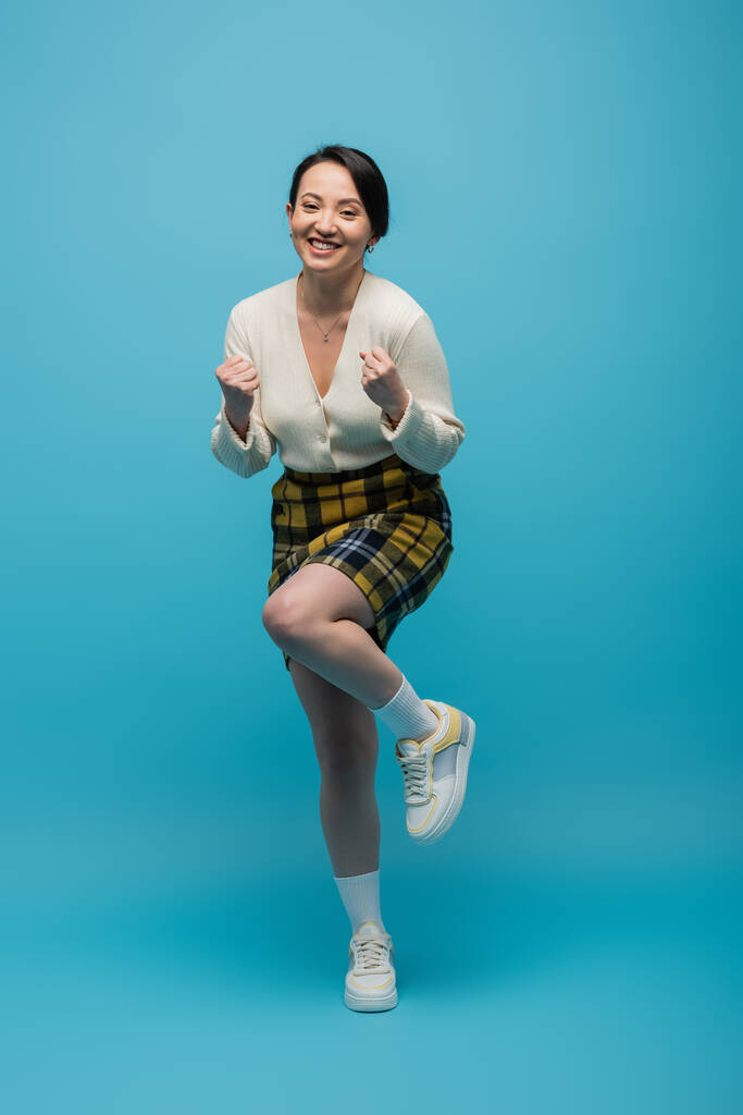 full length of positive asian woman in checkered skirt and sneakers standing on blue  - Foto, imagen