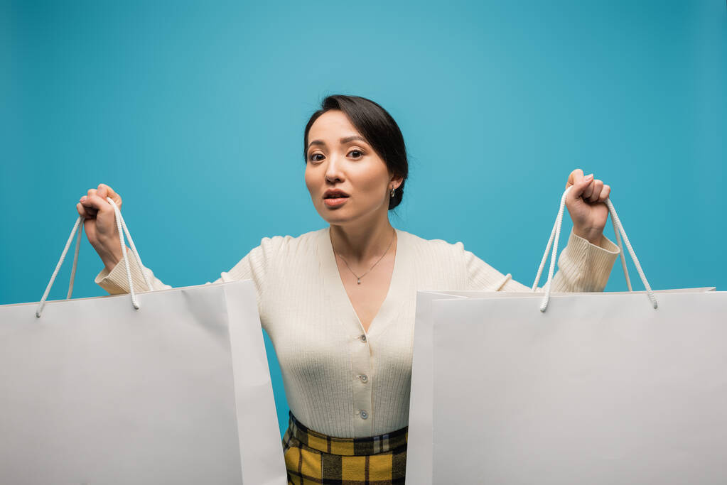 Asian woman holding shopping bags and looking at camera isolated on blue  - Foto, afbeelding