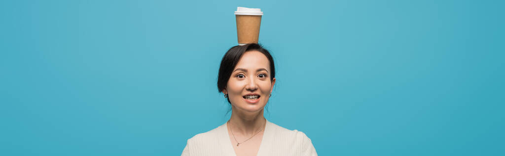 Smiling asian woman holding coffee to go on head isolated on blue, banner  - Valokuva, kuva