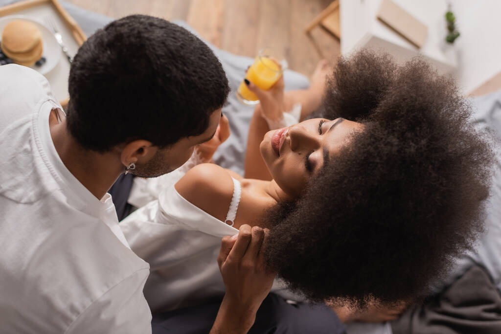 top view of young african american man undressing sexy girlfriend holding blurred orange juice in bedroom - Foto, Imagem