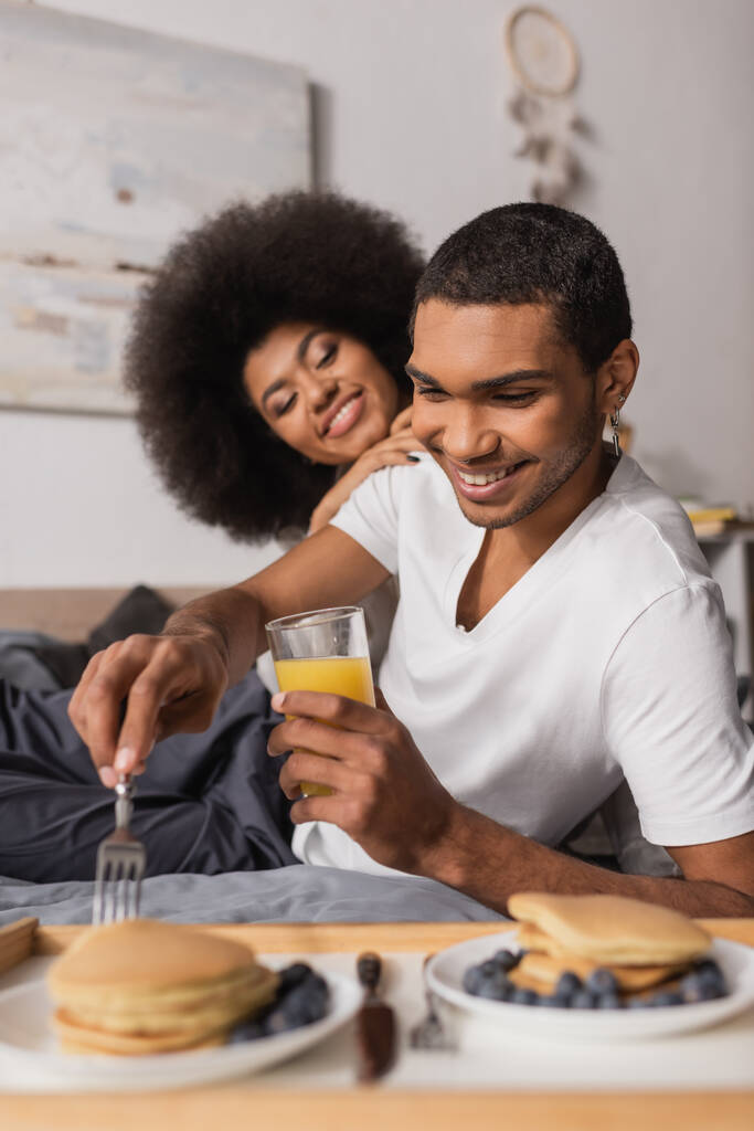 кучерява афроамериканка посміхається біля хлопця, тримає апельсиновий сік і виделку біля розмитих млинців з чорницею
 - Фото, зображення