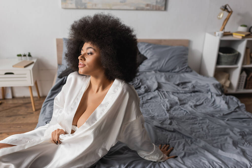 young and sexy african american woman in white robe sitting on bed and looking away - Foto, immagini