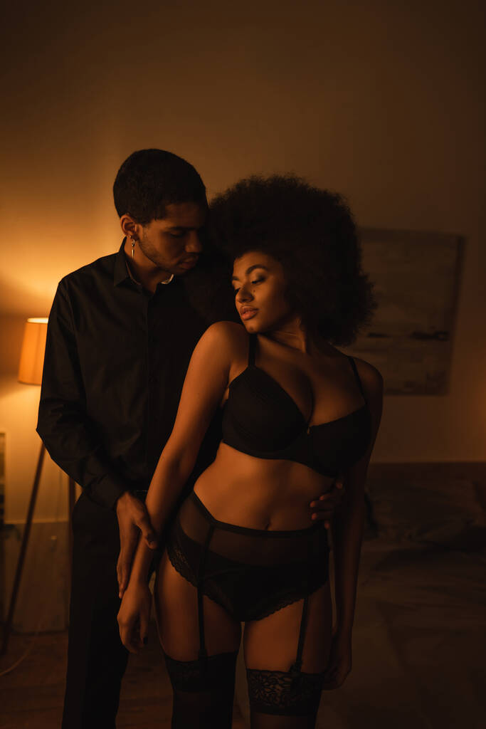 african american man in black shirt near passionate woman in sexy underwear in dark bedroom with lighting - Foto, imagen